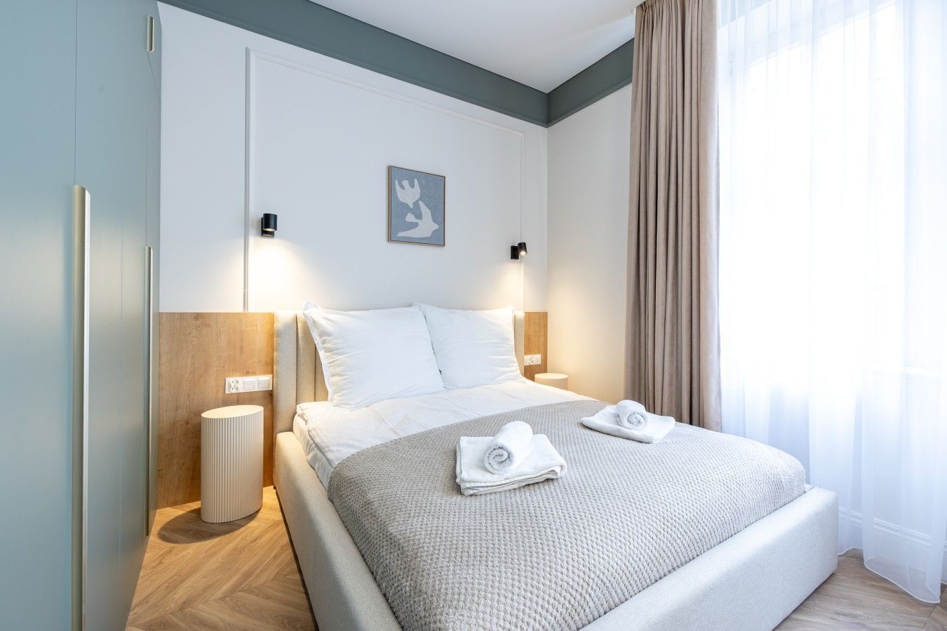 Habitación de hotel con cama, lámparas de noche, mesita de noche, luz en la pared, un cuadro enmarco y ventana con cortinas.