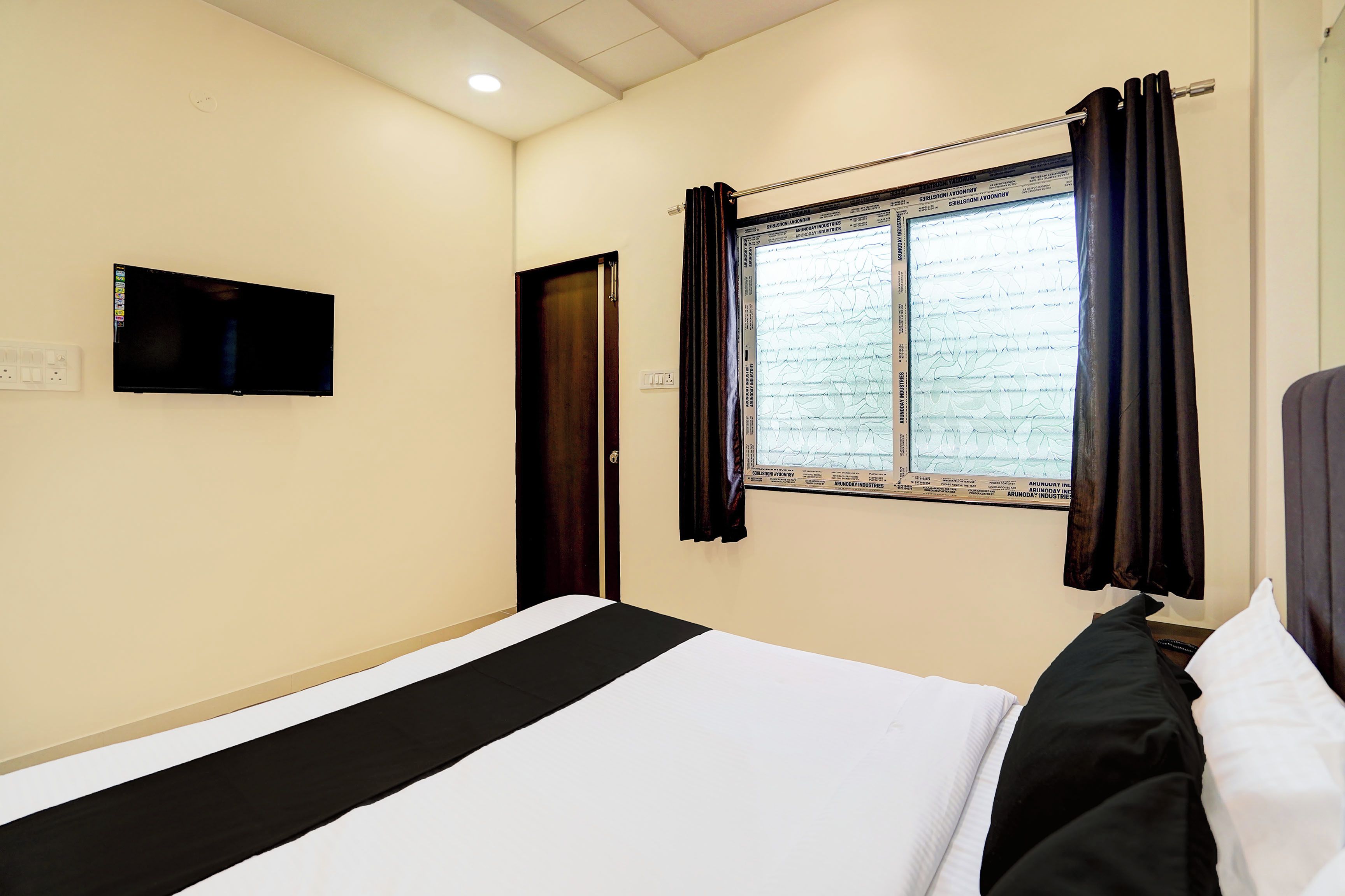 A hotel room with a bed, a flat-screen TV mounted on the wall, a window with curtains, and a door.