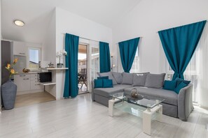 Een moderne woonkamer met een grijze bank, blauwgroene gordijnen, een glazen salontafel en een keukenhoek met een koffieapparaat.