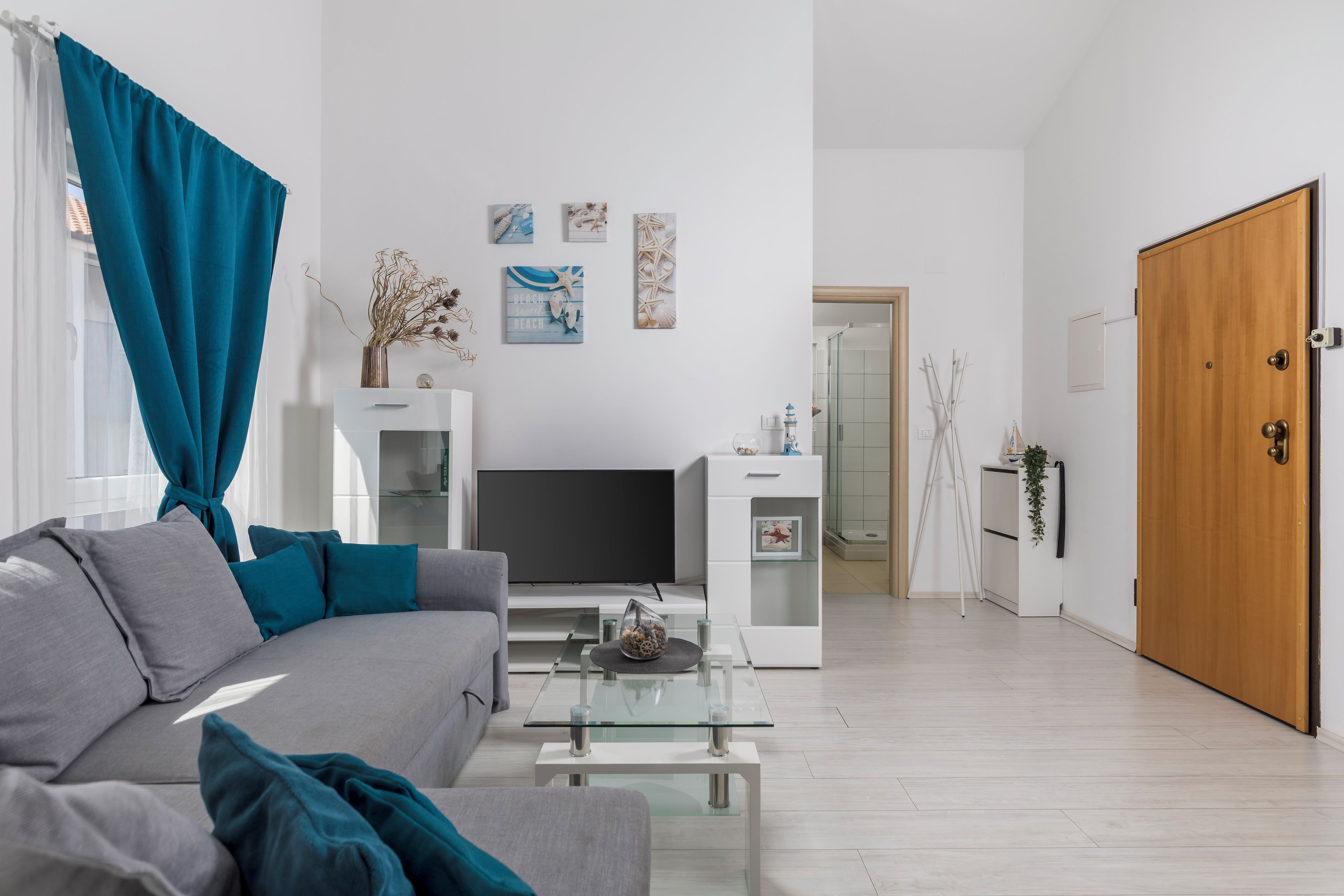 A modern living room with a grey sofa, teal cushions, a glass coffee table, and a flat-screen TV.