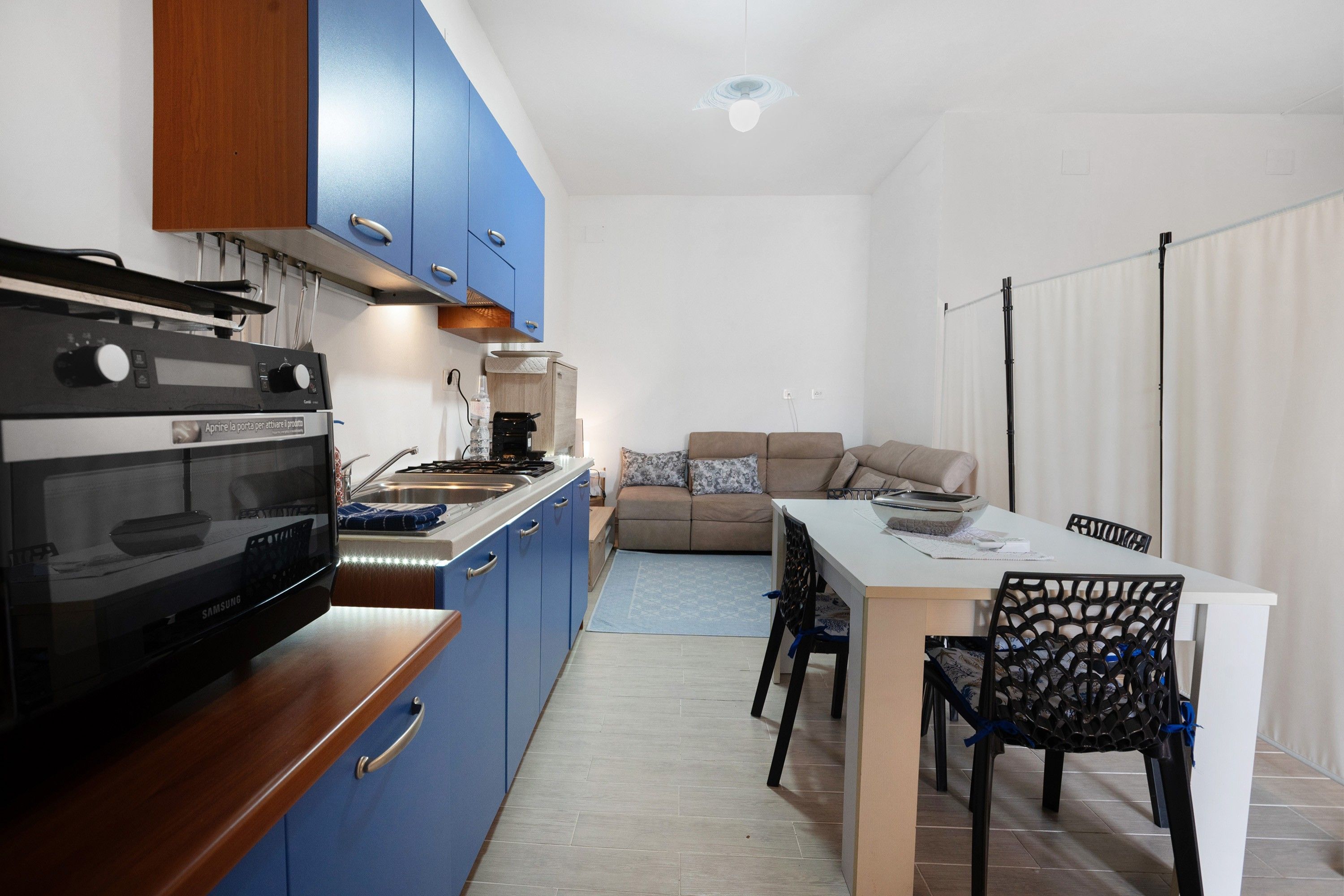 A compact kitchen with blue cabinets, a stove, and a sink. There's a dining area with a table and chairs, and a sofa in the background.