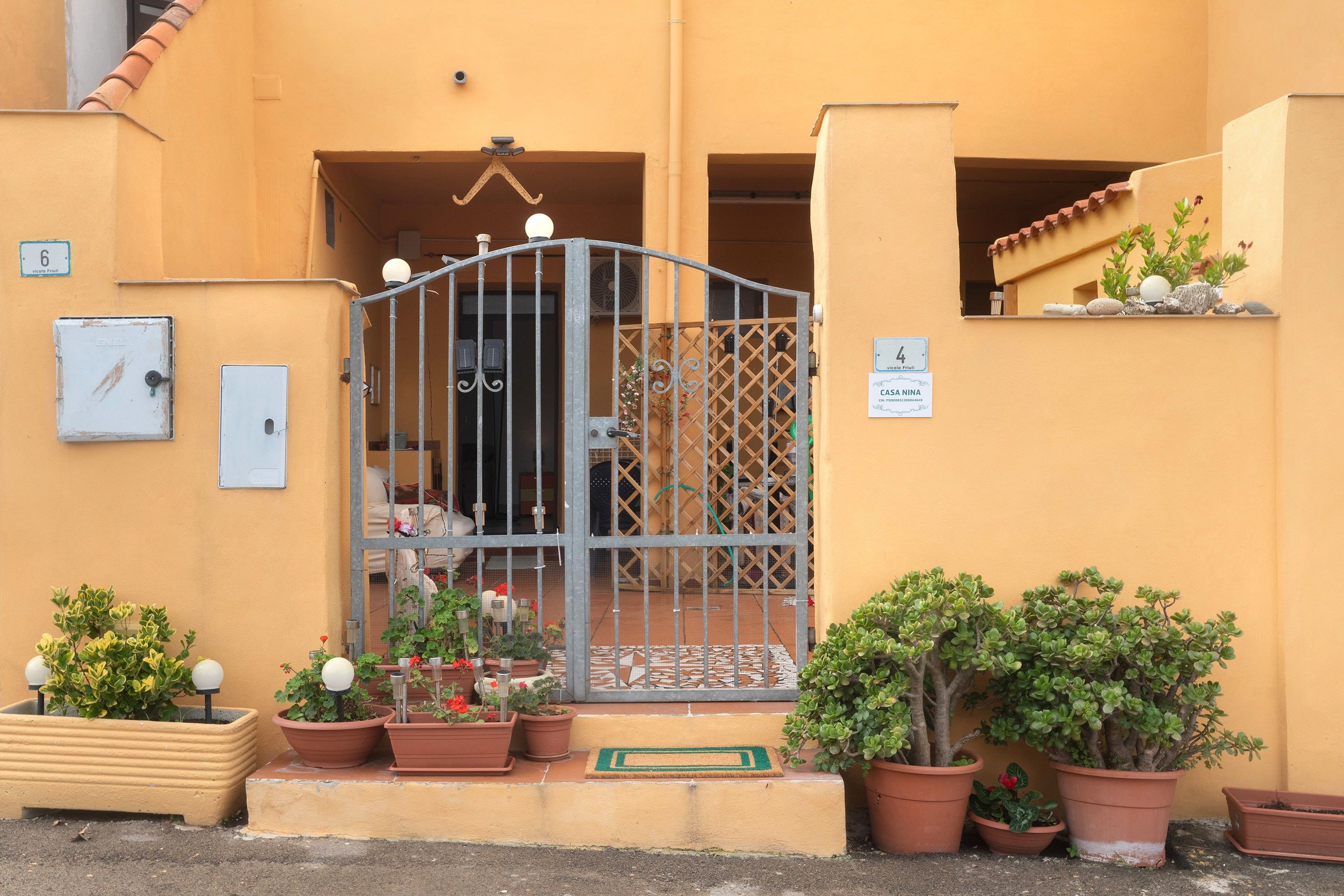 Entrada de um edifício com portão de metal, plantas em vasos e uma placa indicando 'CASA NINA'.