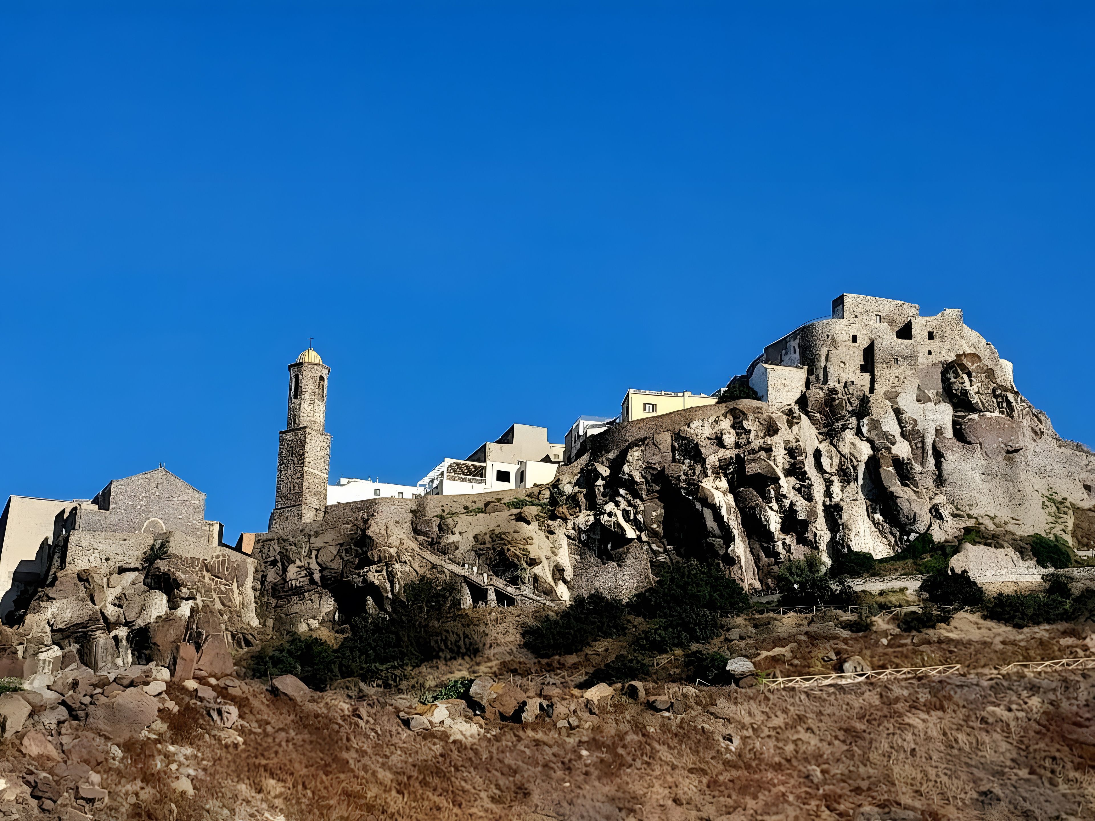 A historic stone building with a prominent tower, situated on a rocky hill with modern buildings in the background.