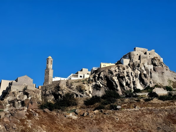A historic stone building with a prominent tower, situated on a rocky hill with modern buildings in the background.