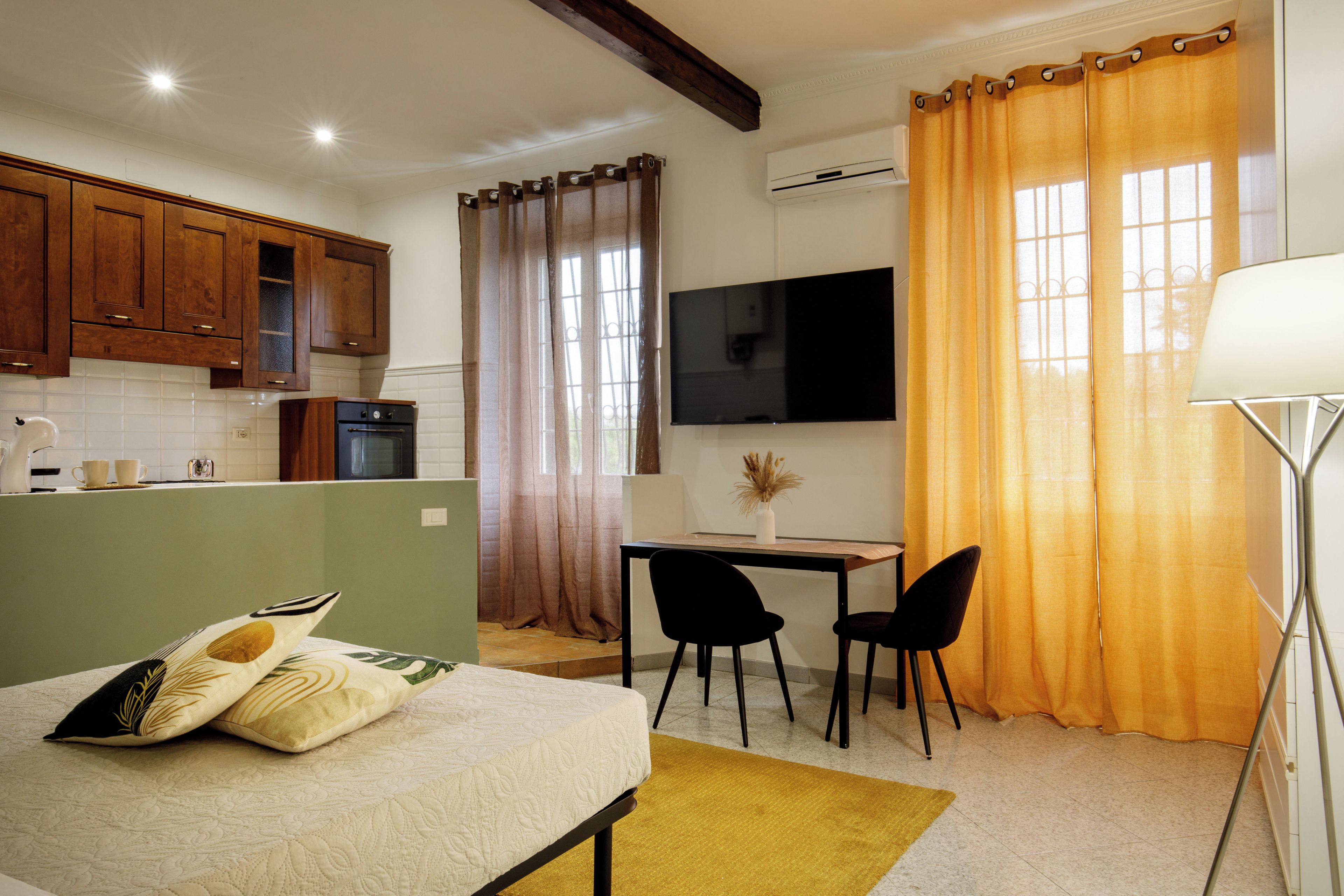 Una camera d'albergo compatta con un letto, una scrivania, due sedie, una TV e un angolo cottura con mobili in legno e un forno a microonde.