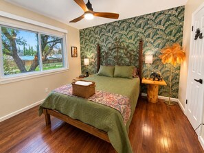 A bedroom with a bed, a wooden floor, a ceiling fan, a window with a view, and a wall with a leafy pattern. - Viva La Vida- Tropical Hacienda + Private Pool (El Paso)
