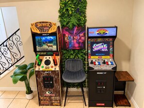 Three arcade machines, a chair, and a small table in a room with a plant and a decorative wall. - Viva La Vida- Tropical Hacienda + Private Pool (El Paso)