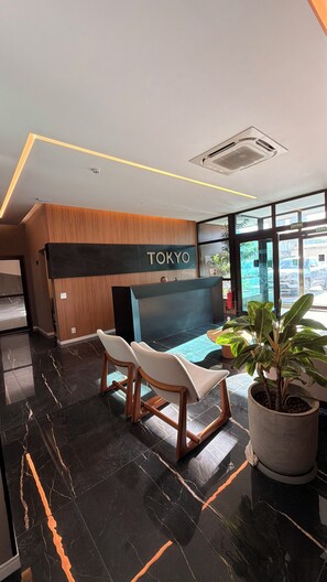 A reception area with a 'TOKYO' sign, a reception desk, two white cushioned chairs, a potted plant, and a view through large windows. - New Listing (Maceió)