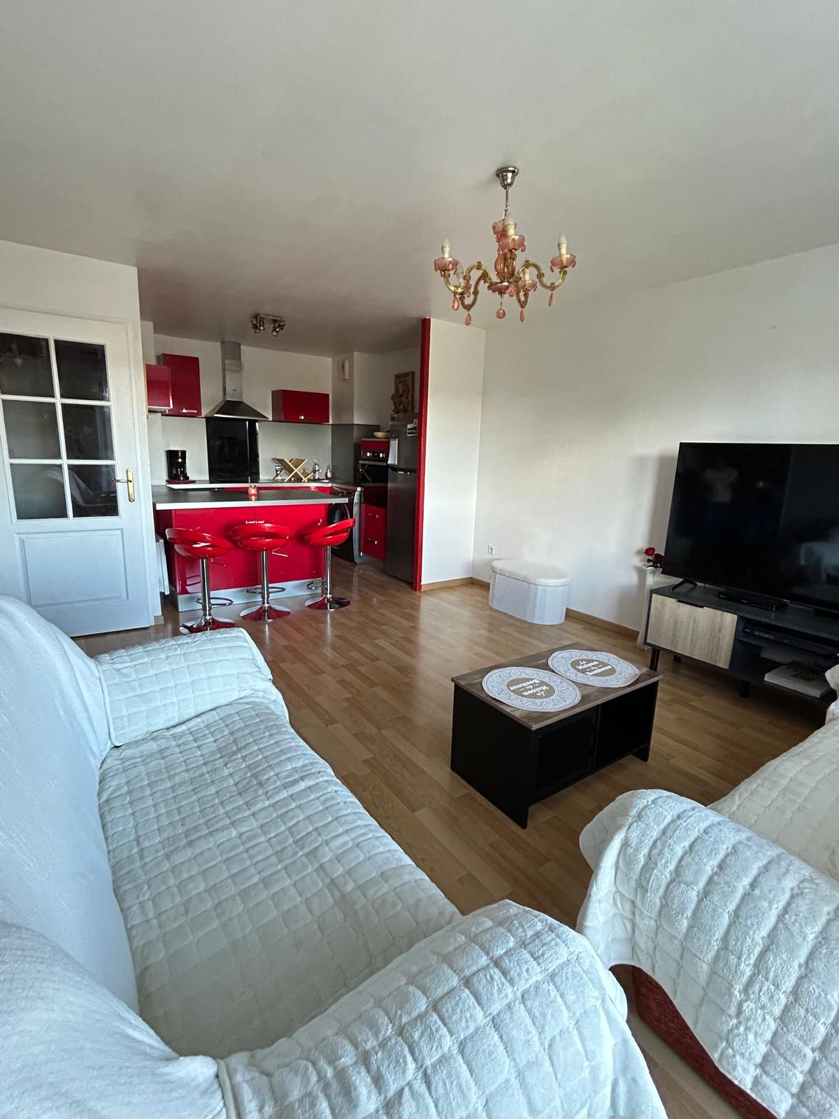 Un salon avec un canapé, une table basse et un téléviseur. Il y a également un coin cuisine avec des meubles de rangement rouges et un lustre.