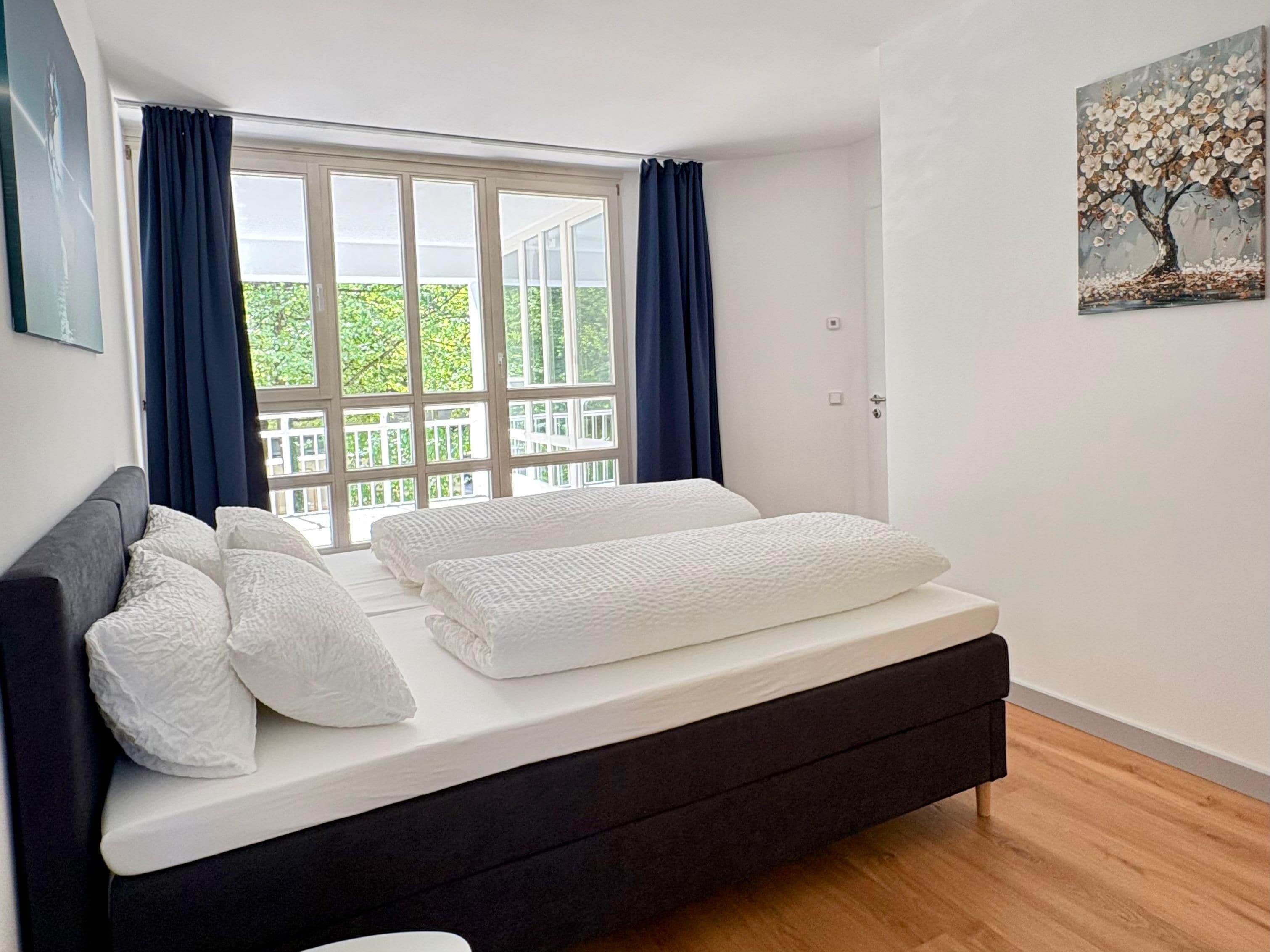 A bedroom with a bed, a window with curtains, a painting on the wall, and a balcony view.