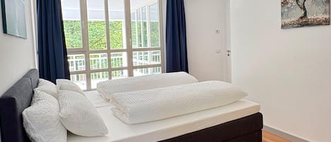 A bedroom with a bed, a window with curtains, a painting on the wall, and a balcony view.