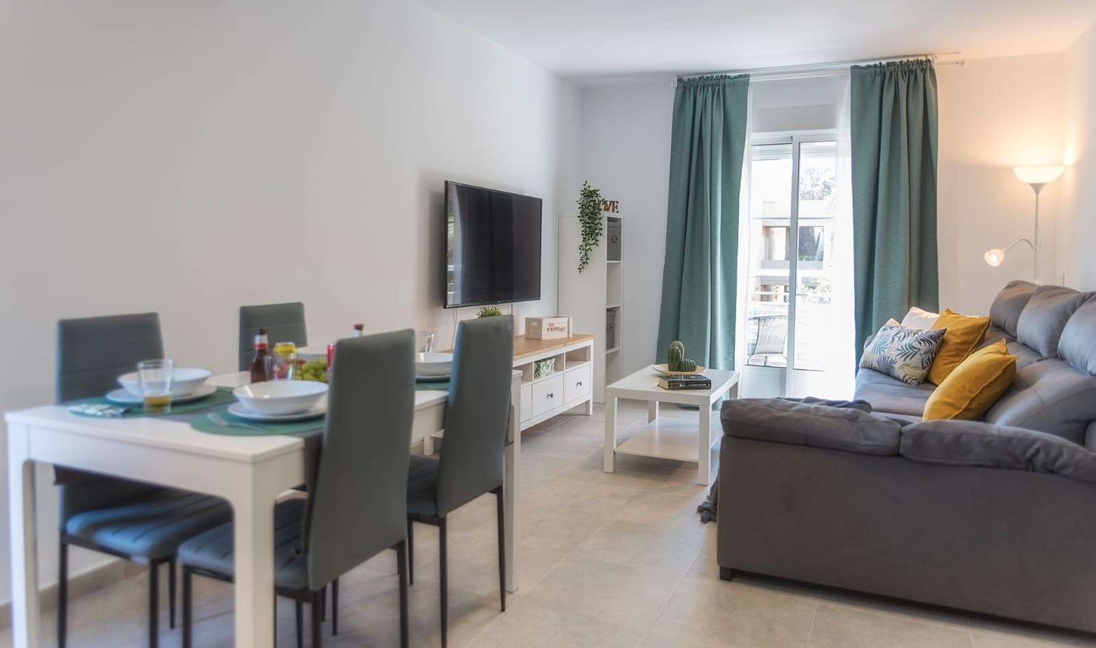 Un salon moderne avec un canapé gris, un coin repas comprenant une table et des chaises, un téléviseur à écran plat fixé au mur et une grande fenêtre avec des rideaux.