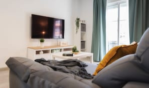 Un soggiorno moderno con un divano grigio, una TV a schermo piatto montata a parete, un tavolino bianco con una pianta e una finestra con tende.