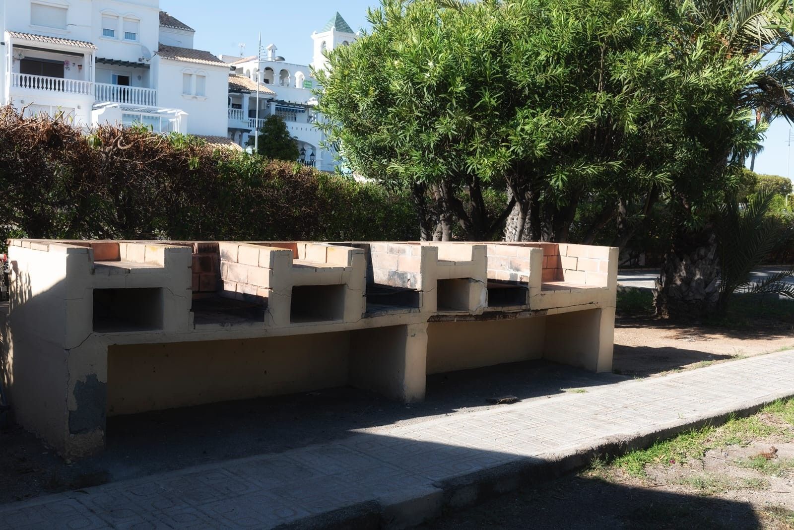 Un banco de hormigón con superficie plana y una parte inferior, ubicado en una zona pavimentada con un árbol y edificios al fondo.