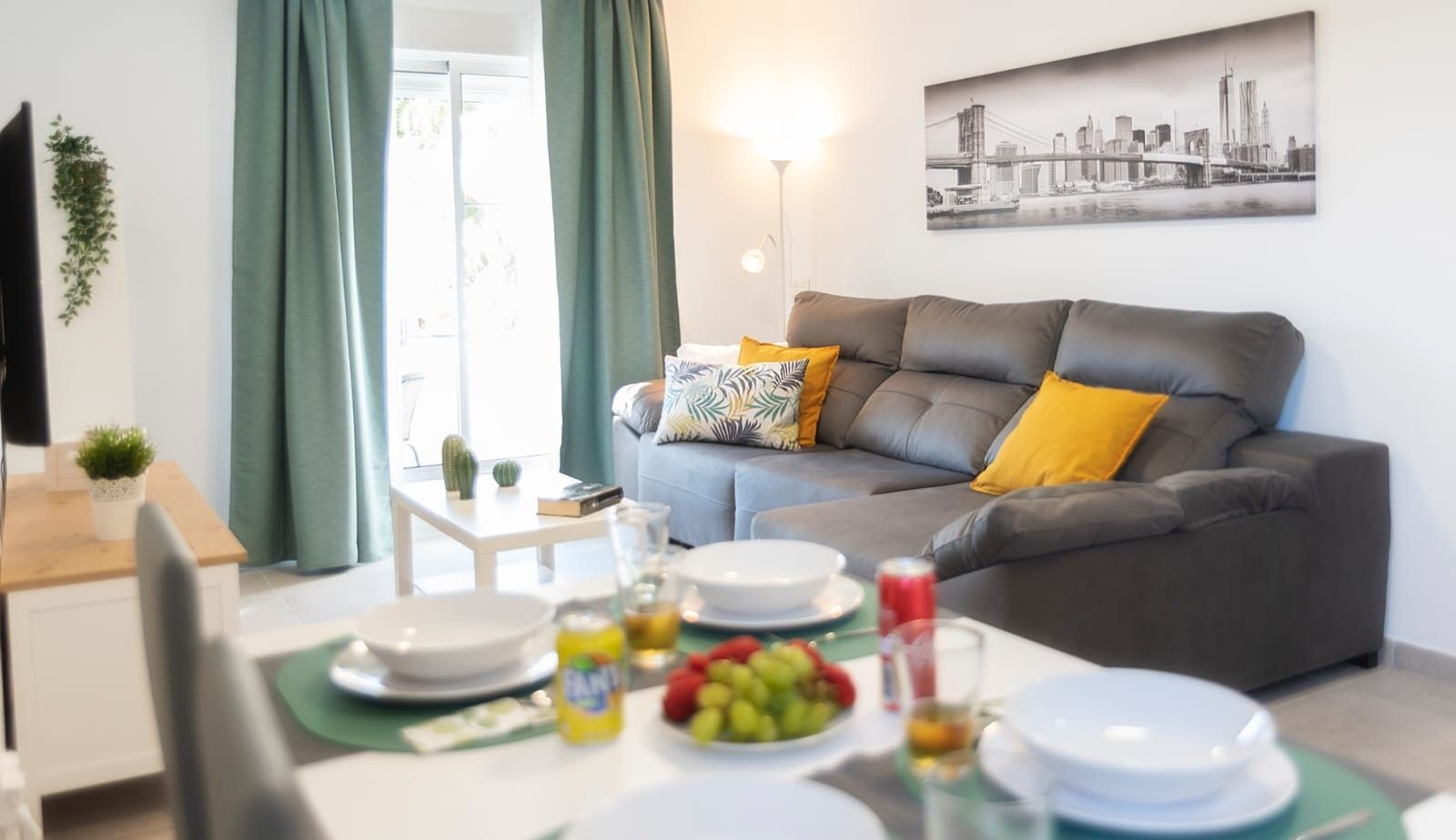 Una sala de estar con un sofá gris, una mesa de comedor dispuesta con platos y un frutero, una mesita con una planta, y un cuadro de un paisaje urbano en la pared.