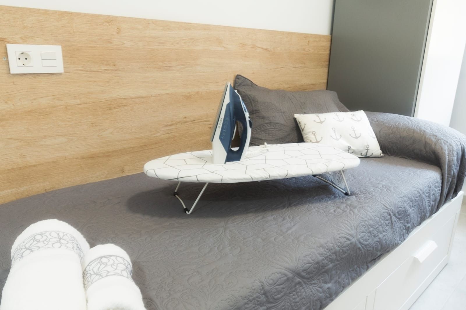 Un lit avec une couvre-lit à motifs gris, un oreiller orné d’imprimés d’ancres et une planche à repasser avec un fer à repasser.