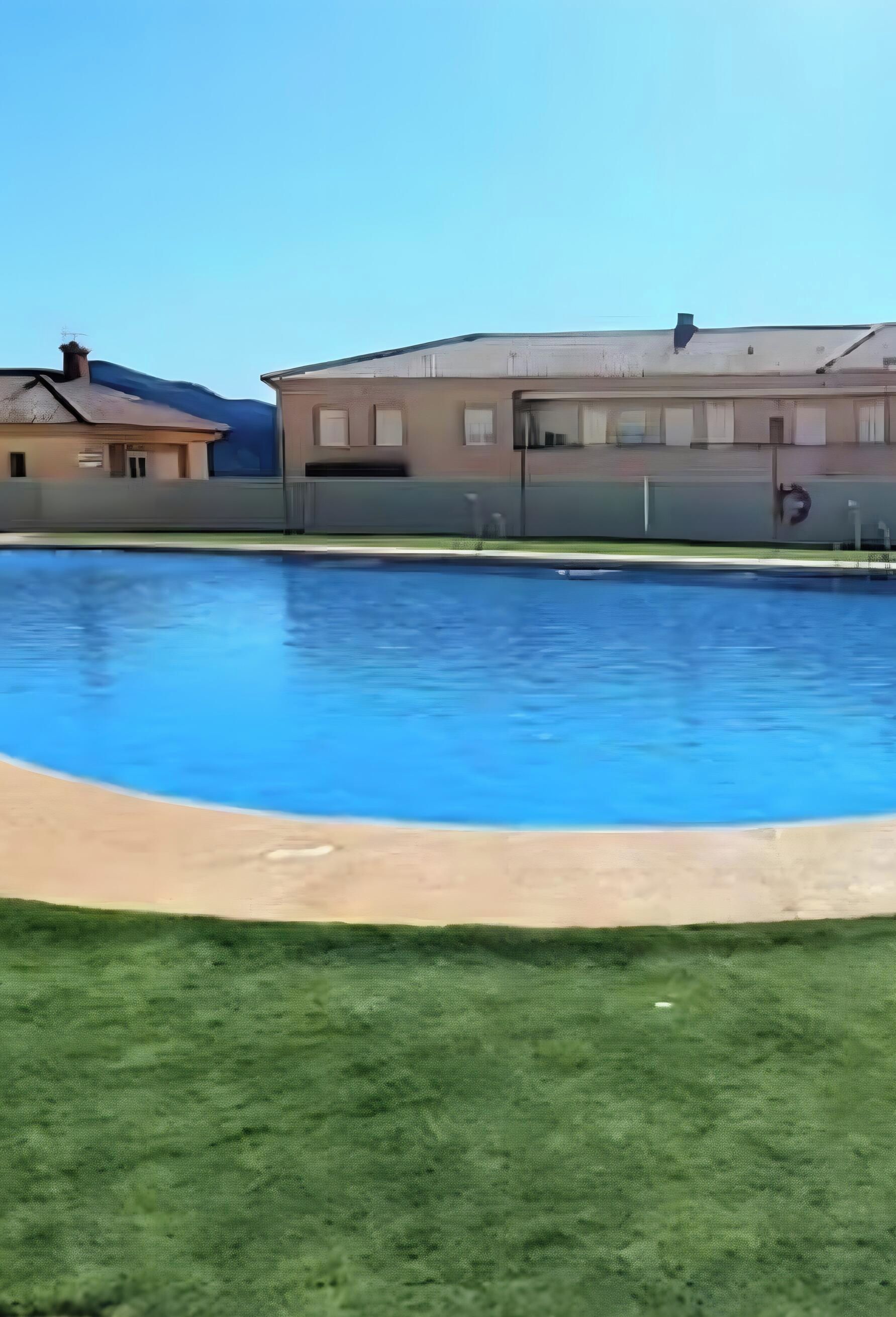 Une piscine avec une pelouse au premier plan et des bâtiments en arrière-plan.
