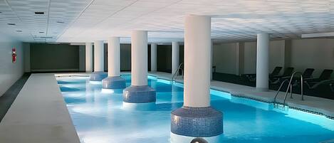 An indoor swimming pool with clear water, surrounded by white pillars and a tiled ceiling.