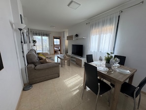 A modern living room with a sofa, TV, dining table set, and a kitchen area.