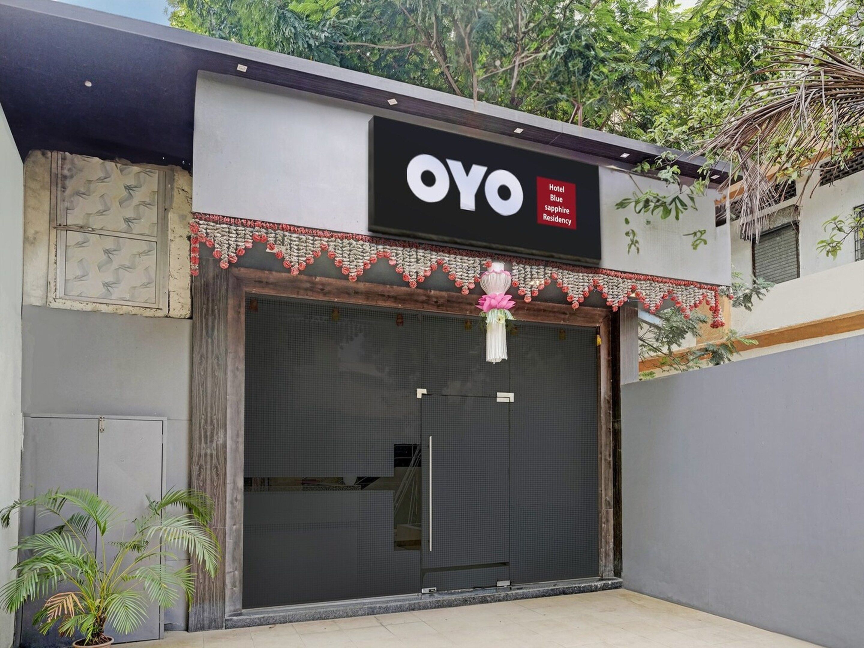 Entrada de un hotel con una persiana baja, un cartel con el nombre 'OYO Hotel Blue Sapphire Residency' y una guirnalda decorativa sobre la entrada.