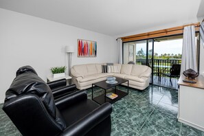 A modern living room with a sectional sofa, a black leather armchair, a coffee table, and a view of the outdoors through a sliding glass door. - Relaxed Island Living Condo Golf Course Views (St.Petersburg)