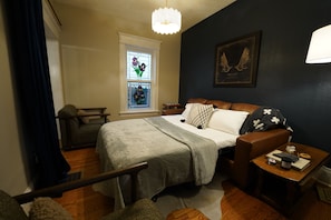 Une chambre à coucher avec un lit, une table de chevet, une fenêtre avec des vitraux, une chaise et un tableau accroché au mur.