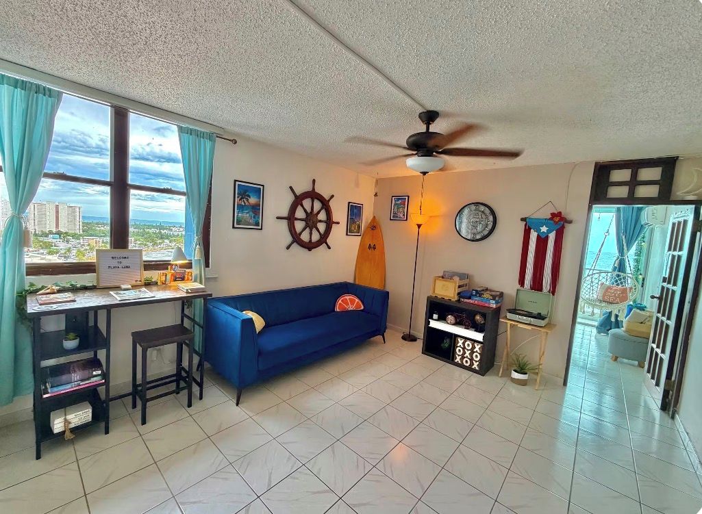 A living room with a desk, a blue sofa, a ceiling fan, and a city view.