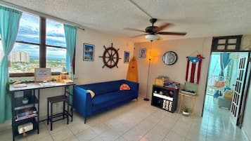A living room with a desk, a blue sofa, a ceiling fan, and a city view.