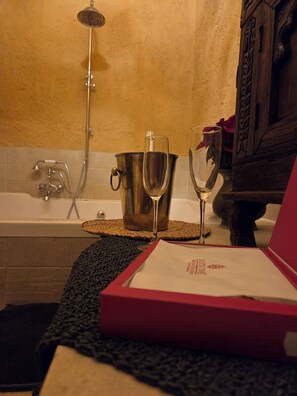 A bathroom with a bathtub, a shower, a metal bucket, a glass of water, a wine glass, and a red box on a textured mat. - New Listing (Orgaz)