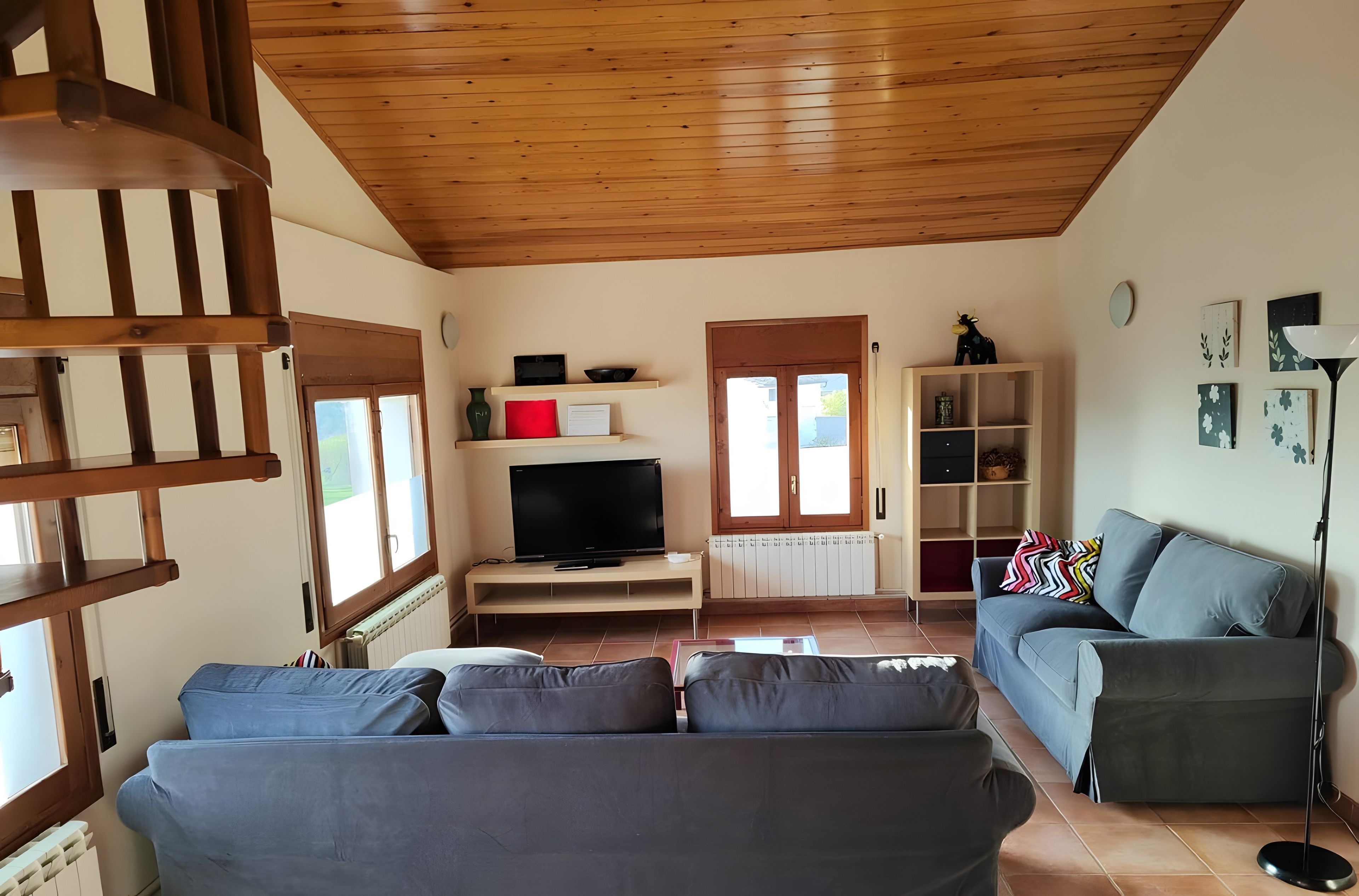 Una sala de estar con techo de madera, televisor sobre un mueble, sofá, aparador y lámpara de pie.
