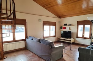 A spacious living room with a wooden ceiling, a grey sofa, a television on a stand, and a staircase.
