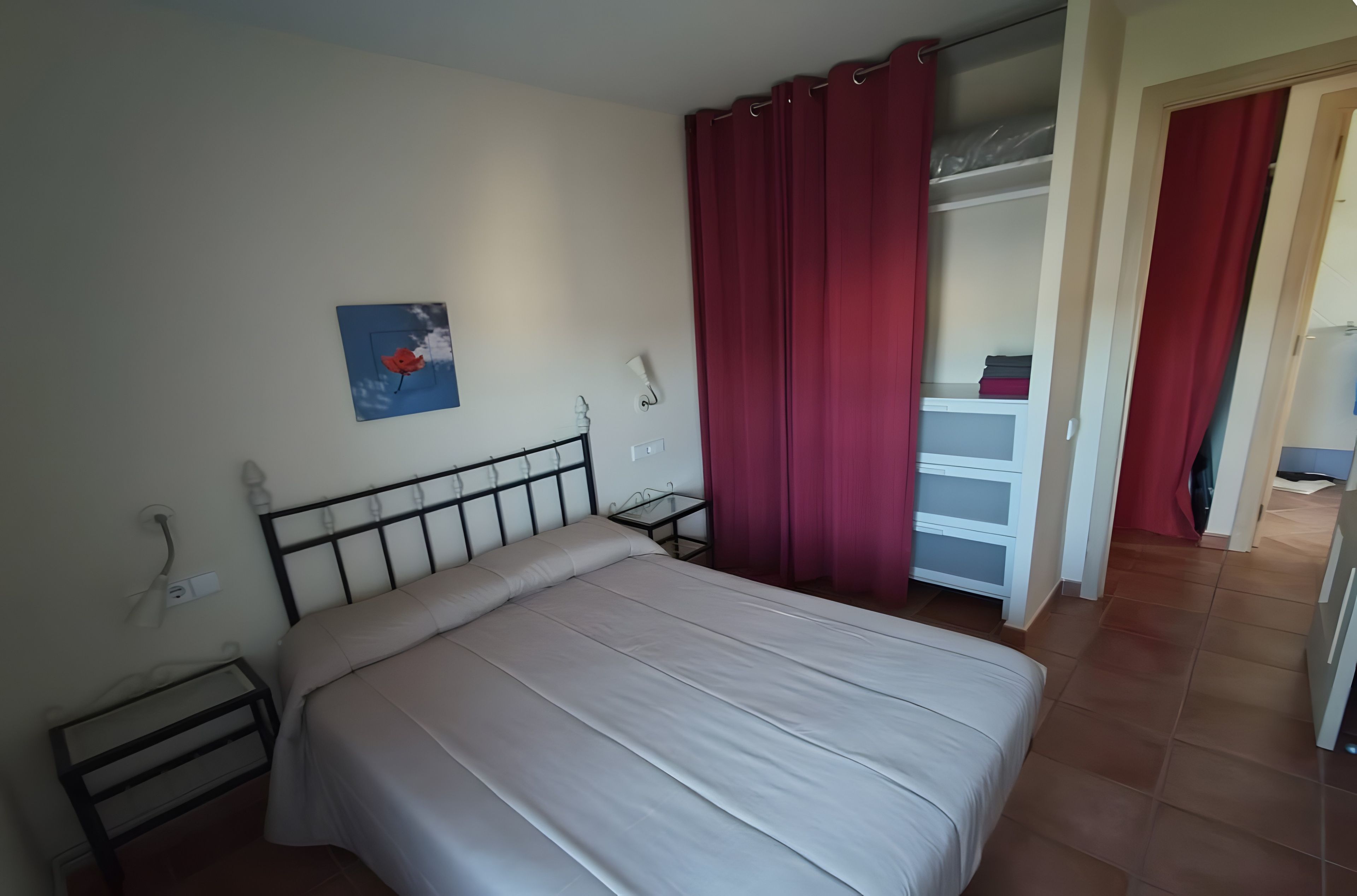 Une chambre à coucher avec un lit, une table de chevet, une lampe fixée au mur, un rideau rouge et un petit tableau.