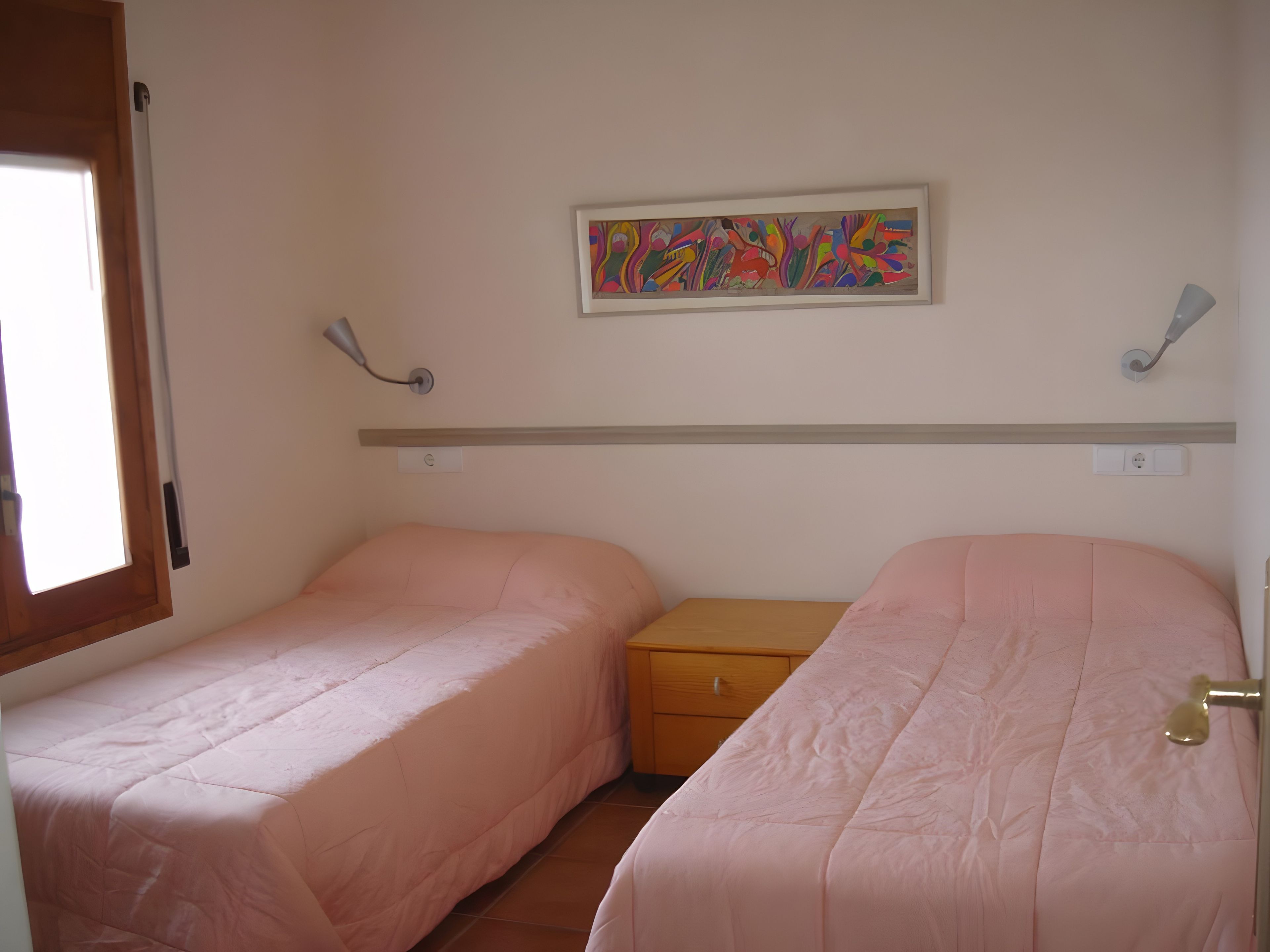 Un dormitorio con dos camas individuales, una mesita de noche, una lámpara de pared y un cuadro abstracto colorido.