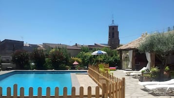 A clear blue swimming pool with a wooden fence, a clock tower, and a sign reading 'TORRES DEL OBISPO' in the background.