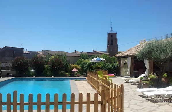 A clear blue swimming pool with a wooden fence, a clock tower, and a sign reading 'TORRES DEL OBISPO' in the background.