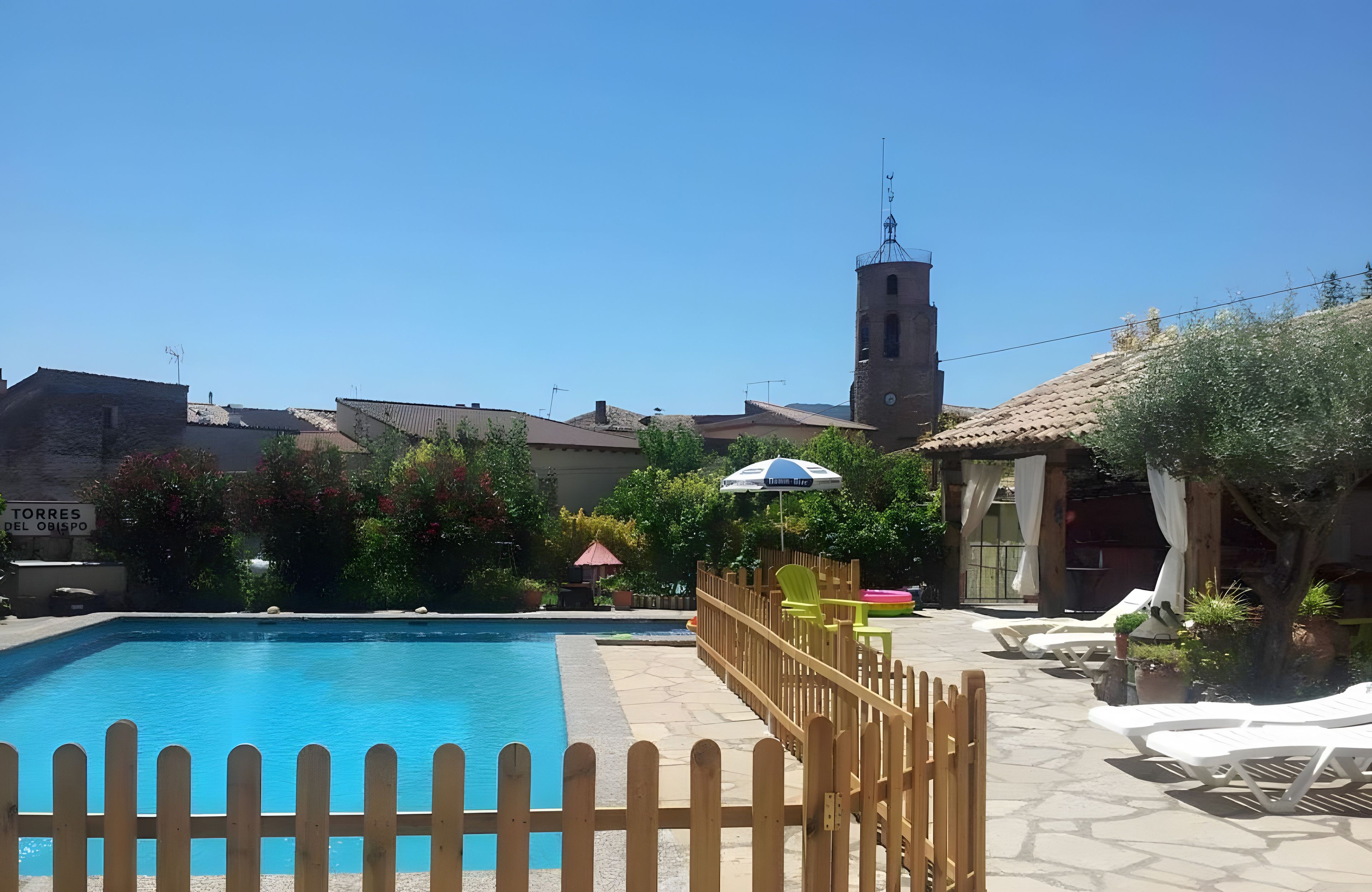 Una piscina azul cristalina con una cerca de madera, una torre del reloj y un letrero que dice 'TORRES DEL OBISPO' al fondo.