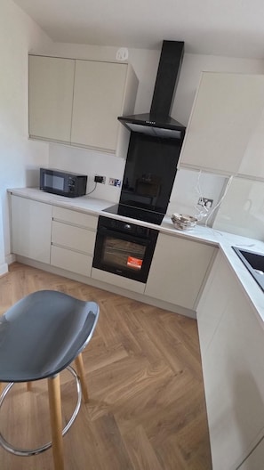 A compact kitchen with a microwave, oven, and stove, a chair, and wooden flooring. - Contractor Stays 4 Bedroom- Free Parking 2 Spaces (Pontefract)