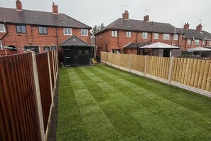 Un jardin à l’arrière d’une maison, avec une pelouse bien entretenue, une clôture en bois et des maisons en arrière-plan.