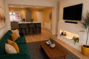 A living room with a green sofa, a wooden coffee table, a fireplace, and a TV mounted on the wall. - Contractor Stays 4 Bedroom- Free Parking 2 Spaces (Pontefract)