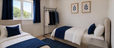 A bedroom with two beds, a window with curtains, a radiator, a clothing rack, and two framed leaf designs on the wall.