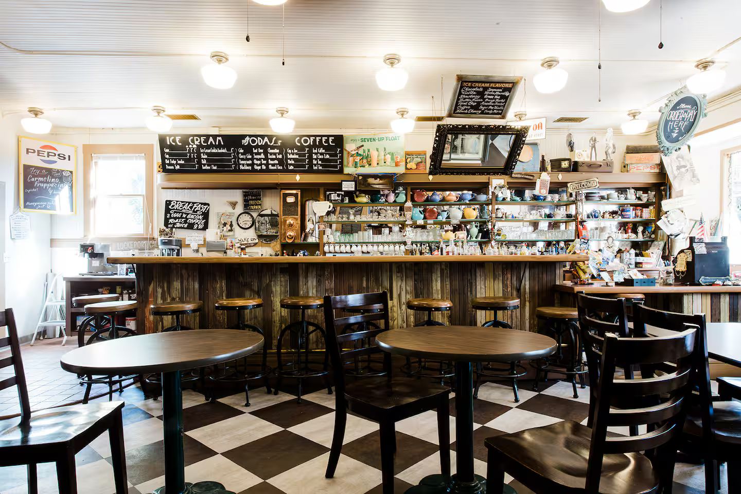 A cafe interior with a bar area, multiple seating options including tables and stools, and a variety of items displayed on shelves behind the bar.