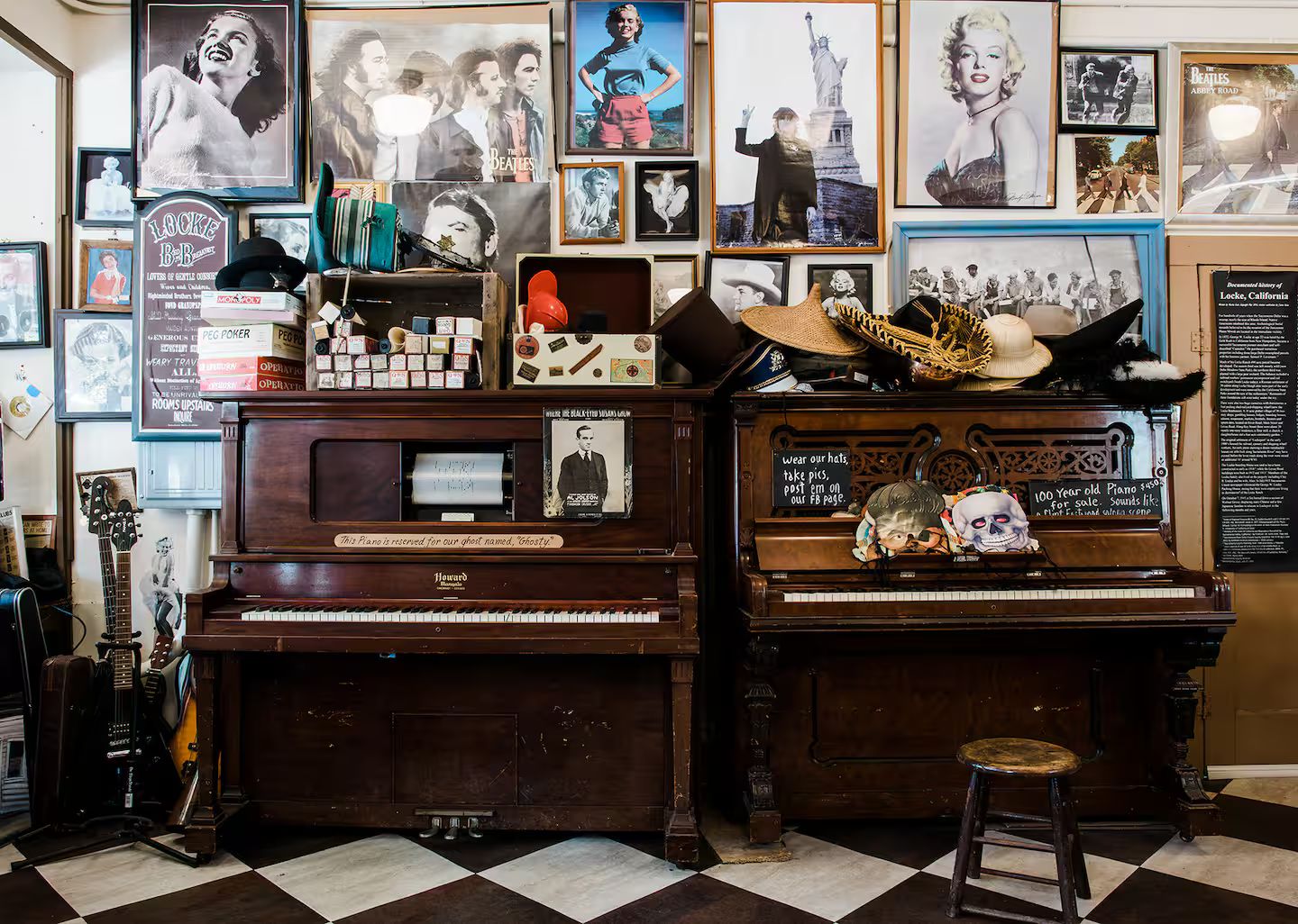 A room with two pianos, one of which is labeled 'Howard.' There are various posters and memorabilia on the walls, including a framed picture of a man with a hat and a sign that reads '100 Year old Piano.' A stool is placed in front of the right piano.