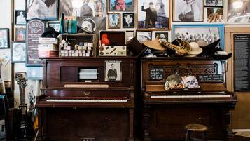A room with two pianos, one of which is labeled 'Howard.' There are various posters and memorabilia on the walls, including a framed picture of a man with a hat and a sign that reads '100 Year old Piano.' A stool is placed in front of the right piano.