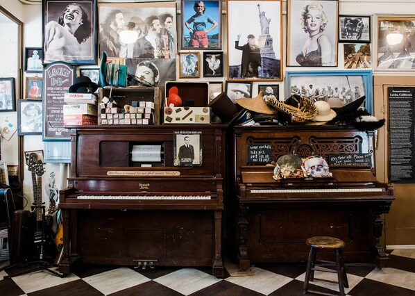 A room with two pianos, one of which is labeled 'Howard.' There are various posters and memorabilia on the walls, including a framed picture of a man with a hat and a sign that reads '100 Year old Piano.' A stool is placed in front of the right piano.
