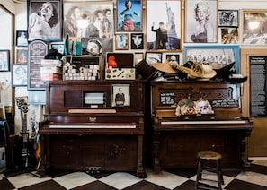A room with two pianos, one of which is labeled 'Howard.' There are various posters and memorabilia on the walls, including a framed picture of a man with a hat and a sign that reads '100 Year old Piano.' A stool is placed in front of the right piano.