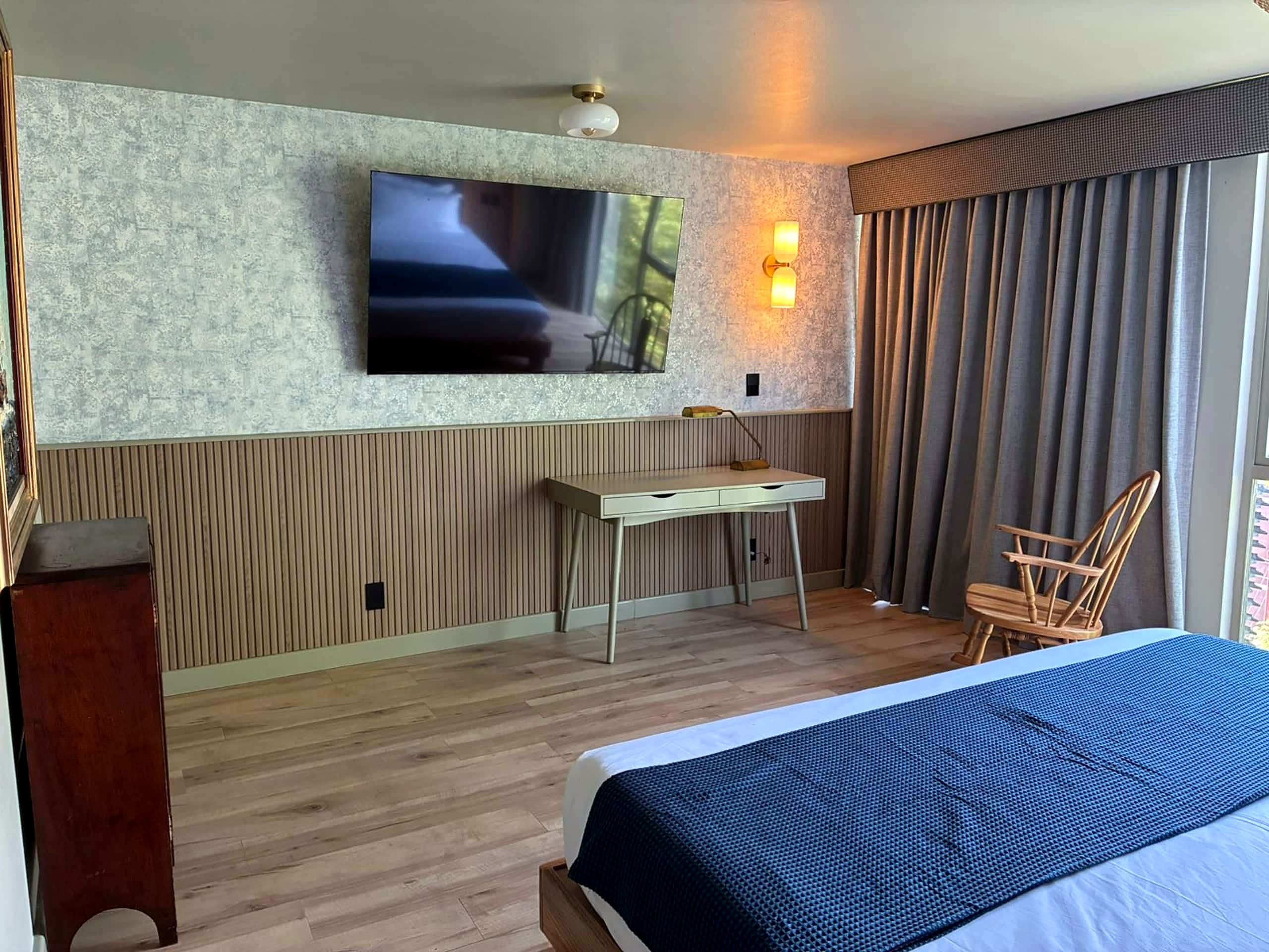 Une chambre d’hôtel comprenant un lit, un bureau, une chaise, un téléviseur fixé au mur et une fenêtre avec des rideaux.