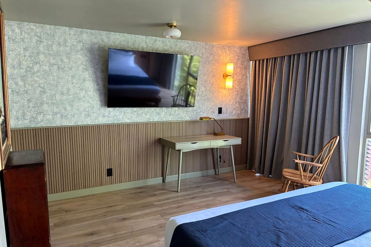 Habitación de hotel con cama, escritorio, silla, televisión montada en la pared y ventana con cortinas.
