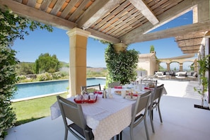 Zona d'exterior per dinar amb vista a la piscina, coberta per una pèrgola de fusta, i envoltada de vegetació.