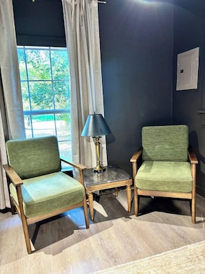 Twee groene fauteuils met houten frames, een klein tafeltje met een lamp, en een raam met gordijnen.