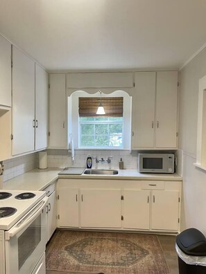 A compact kitchen with white cabinets, a microwave, a stove, and a window with blinds. - Davis St Apartment Complex | Whole House Near SFA (Nacogdoches)